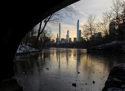 A serene sunset illuminates the New York skyline, seen across a frozen pond from under a bridge in