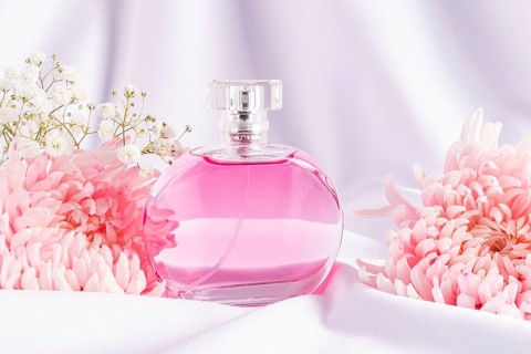 A round glass perfume bottle filled with pink liquid, surrounded by delicate pink and white flowers