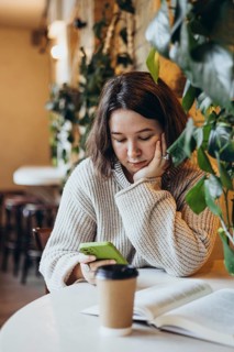 A young woman in a sweater reads a book at a table in a bookstore Surrounded by plants, she holds a