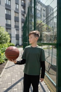 A young boy stands on a paved path in an urban setting, holding a basketball in one hand. He is