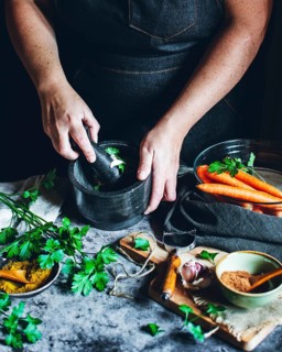 Hands crush fresh herbs in a mortar, surrounded by vibrant vegetables and spices. The scene