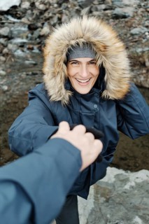 A joyful woman in a warm winter jacket with a fur-lined hood reaches for a helping hand while
