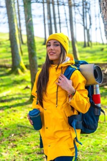 Hiker woman with yellow raincoat and backpack walking in forest, enjoying trekking and drinking hot