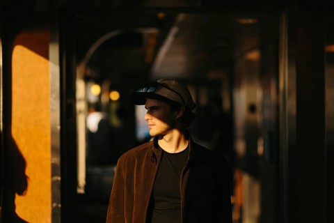 A man stands in warm, moody sunlight, wearing a cap and sunglasses in New York. The indoor setting
