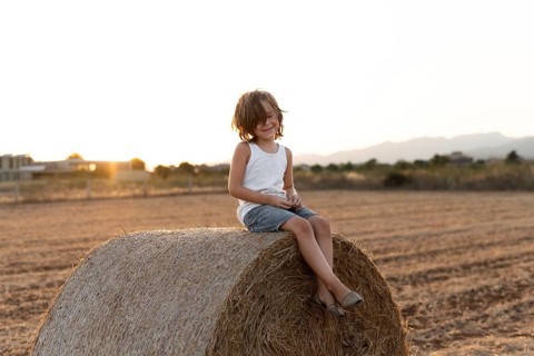 A young child sits on a hay bale in a field at sunset, enjoying the serene rural landscape. The