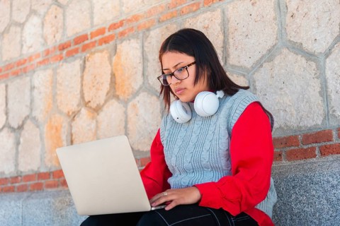 A young Latina wearing glasses and headphones around her neck sits against a stone wall, focused on