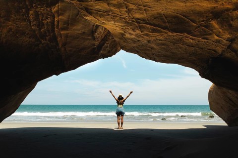 A young woman stands in a cave with arms raised, facing the ocean at Mancora, Peru. The image
