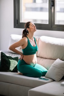 A pregnant woman in a green yoga outfit kneels on a sofa in a sunlit room, taking a moment to relax