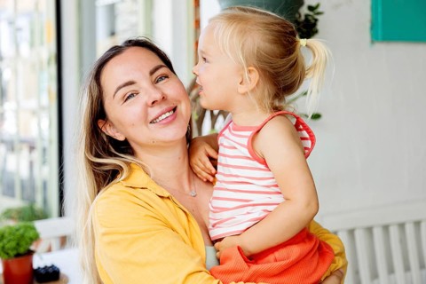 A smiling mother holds her young daughter in her arms while enjoying a sunny day outdoors. The