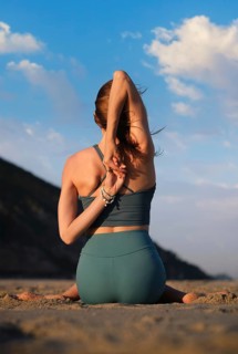 A woman gracefully sits in Gomukhasana, performing yoga by the beach during a stunning sunset,