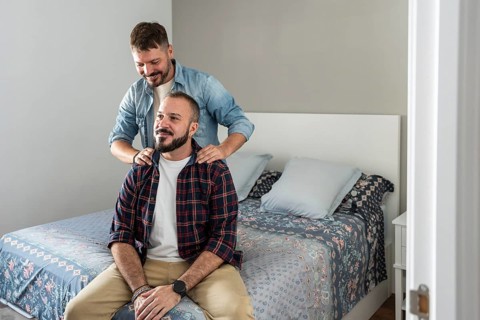 A joyful gay couple sharing a tender moment in their bedroom. One partner stands behind, gently