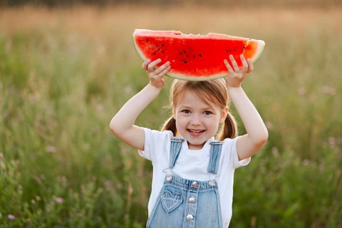 A smiling child holds up a slice of juicy watermelon in a lush green field, capturing the essence