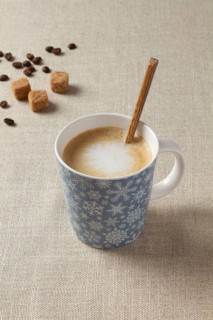 A warm cup of coffee with milk, resting on a beige fabric surface. The mug features a festive