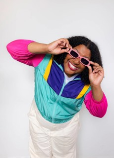 A joyful american woman wearing a vibrant windbreaker and pink sunglasses poses playfully against a