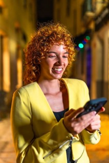 Vertical portrait of a happy woman with curly hair using phone in the city at night