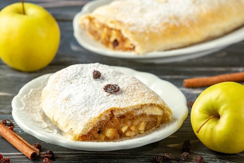 Apple strudel on plate with powdered sugar and raisins, next to yellow apples and cinnamon sticks