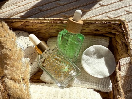 A wicker basket holds two dropper cosmetic bottles, white cotton pads, and natural elements, all