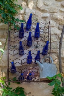 A wall shelf with blue bottles and an old watering can in front of a stone wall. Plants protrude