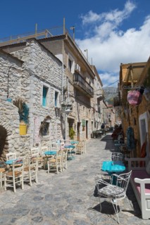 A sunny alleyway view with cafés and traditional stone buildings under a partly cloudy blue sky,