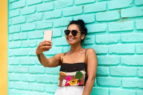 A young Latin woman smiles broadly while taking a selfie against a vibrant turquoise brick wall.