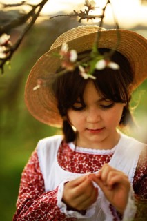 A young girl in a straw hat and floral dress gently examines a flower. The scene captures a serene