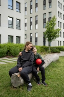 A mother and her young son sitting on a log outdoors, sharing a joyful moment. The boy holds a red