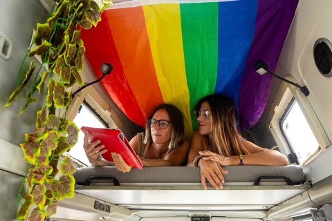 Two women relax in a camper van, using a tablet under a colorful rainbow flag. The vibrant scene