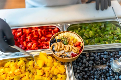 Chef in black gloves preparing an acai smoothie bowl, adding mango with a spoon from a buffet of