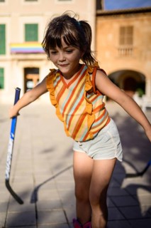 A young girl enjoys playing street hockey, wearing a colorful striped top and shorts. The sunny