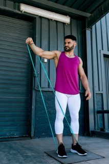 A fit man performs a shoulder exercise using a resistance band outdoors. Wearing athletic attire,