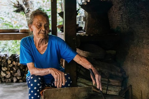 Elderly woman in a blue shirt sits next to a stack of wood and cooking balinese coffee, exuding