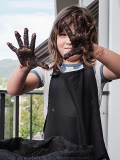 A young girl with messy soil covered hands proudly displays her dirty hands to the camera,