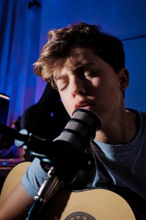 A young musician, sings into a microphone while playing an acoustic guitar in home studio. The blue