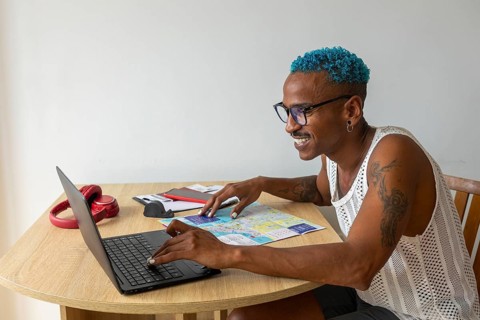 A smiling LGBTQIA+ individual with blue hair plans holidays using a laptop and map, embracing