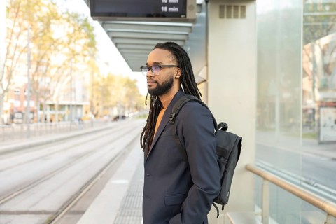 A cuban businessman with long dreadlocks and glasses waits at a tram stop in a bustling city.