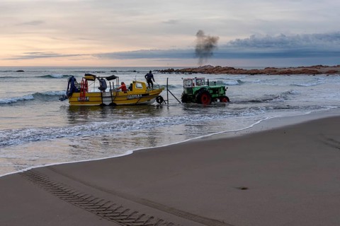Yellow fishing boat with crew is pulled from sandy beach into water by green tractor, rocks in the