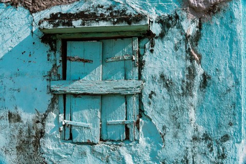 Close-up of a weathered light blue wall with a barred wooden window, reflecting the unique