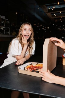 Woman excitedly opening a pizza box at a table, with dim restaurant ambiance. Perfect for visuals