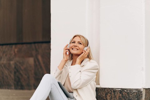 A businesswoman in casual attire sits against a building, wearing headphones and enjoying music.
