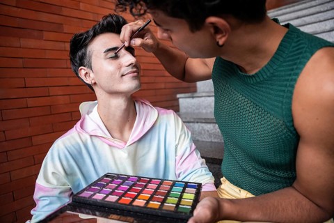 A gay couple shares a joyful moment as one applies makeup on the other using a vibrant colorful