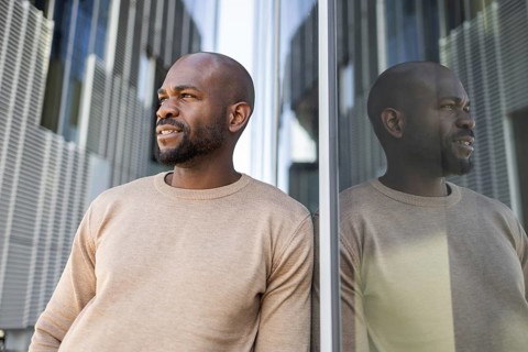 A thoughtful cuban man in a beige sweater stands beside a reflective glass wall, with modern