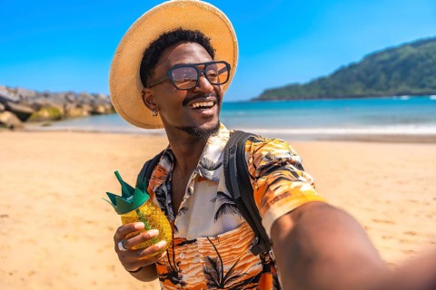 Young black man enjoying summer vacation taking selfies on a tropical beach while holding a