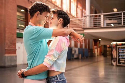 A joyful gay couple embracing closely in a sunlit indoor space. They share a tender moment, smiling