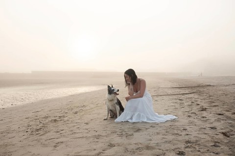 A woman in a flowing white dress sits on a sandy beach with her dog during a tranquil sunrise The