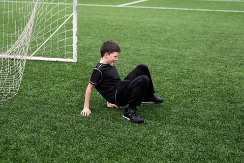A boy dressed in black sportswear smiles on green artificial turf next to a soccer goal, capturing