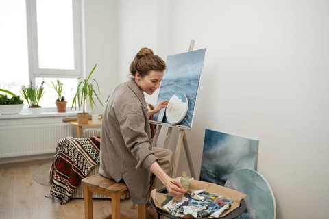 An artist in a cozy, light filled home studio painting a seascape. Surrounded by plants and art