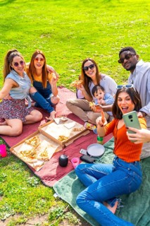 Happy multi ethnic friends eating pizza and taking selfies during a sunny picnic in a park