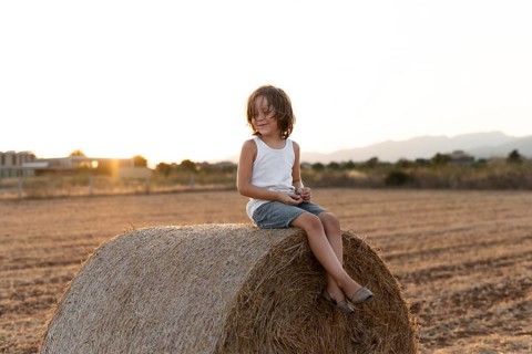 A child in casual attire sits on a large hay bale in a field. The warm sunset in the background
