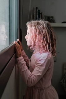 A young girl with curly hair gazes out a window, her reflection visible. She wears a pink dress,