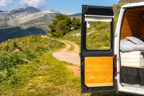 Open door of a camper van reveals a scenic Greek landscape with winding paths and lush greenery,